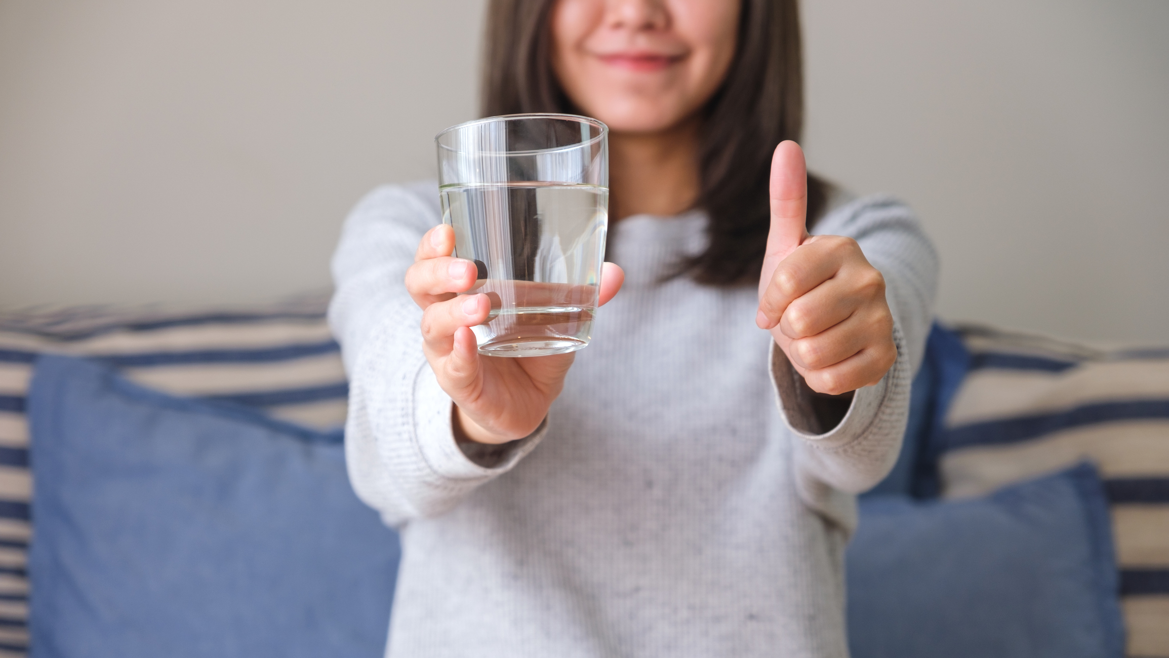 Closeup image of a young woman making thumb up hand sign while holding a glass of water